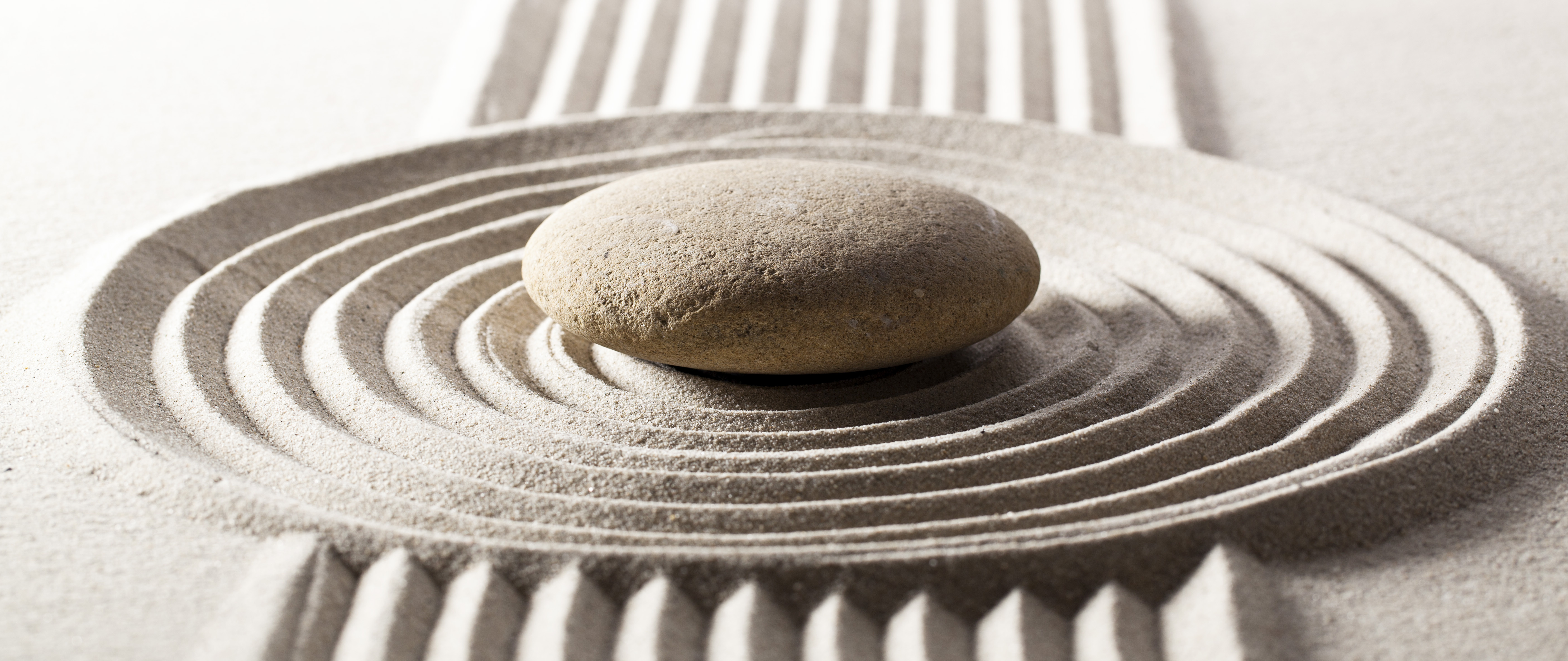 Un giardino zen minimalista con sabbia rastrellata in motivi concentrici attorno a una pietra levigata. Palizzate in legno leggero sono visibili sullo sfondo, creando un'atmosfera di armonia e serenità. La luce morbida illumina i dettagli della sabbia, evocando il concetto di ordine sacro e meditazione nel paesaggio.